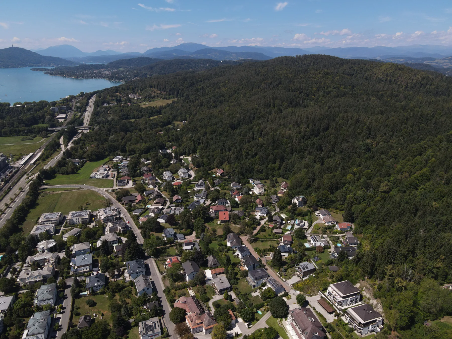 Drohnenaufnahme des grünen Klagenfurter Wohnumfelds mit Wörthersee und umliegender Berglandschaft; der zukünftige Standort von AURA ist markiert – mit KI bearbeitet